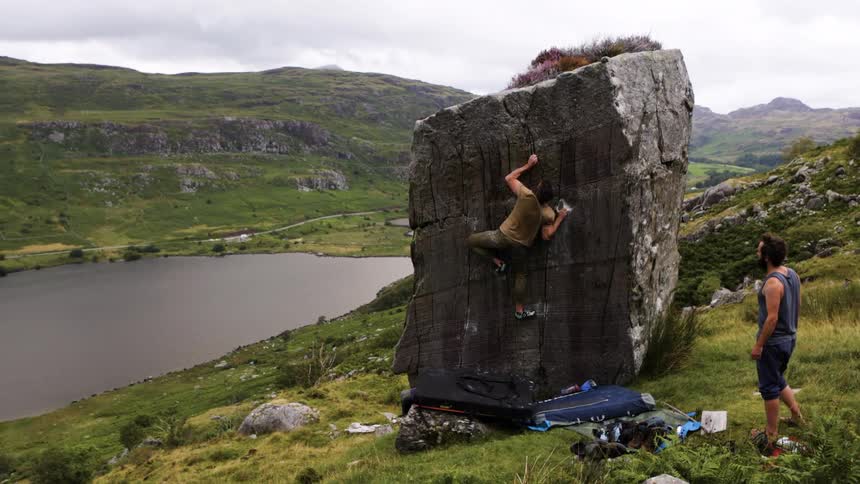 A young man climbing a large rock outdoors on a sloppy terrain during daytime