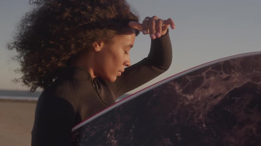 A woman with big curly hair wearing a wet suit stands on a beach with a surfboard under her arm looking at the sea while keeping her hand over her forehead as protection from the wind and the sun while a dog passes her by