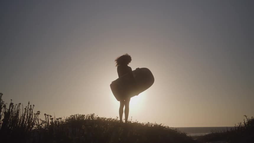 A person with big curly hair standing on a beach dune with a short board under their arm looking at the ocean