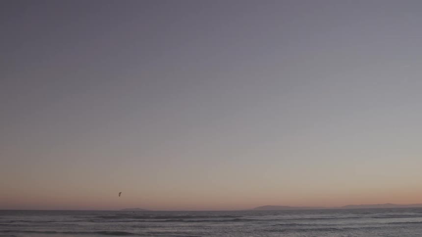 A wide view shows the gentle waves of the ocean rolling onto shore beneath a hazy soft pastelcolored sky at dusk with a distant coastline silhouette on the horizon and a small bird flying in the distance