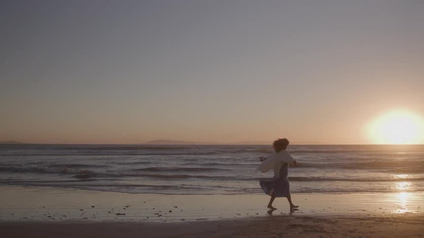 A silhouetted person wearing a light dress walks slowly barefoot across the wet sand of a beach with ocean waves and a bright golden sunset over a distant hazy coastline
