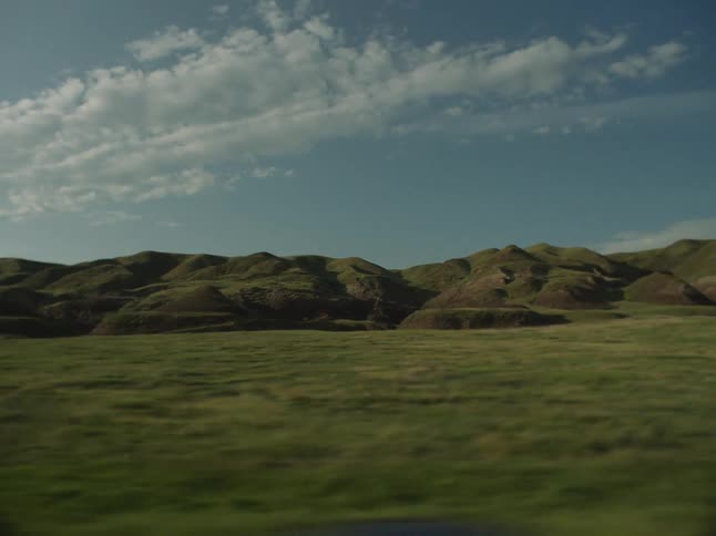 A wideangle side view captures a rapid movement across a flat green grassy plain with the background consisting of a range of rounded brown and green hills under a blue sky with scattered white clouds