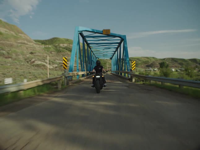 A person riding a dark motorcycle drives toward the camera crossing a wooden bridge with a blue metal truss structure then exits the bridge onto a paved road under a bright blue sky