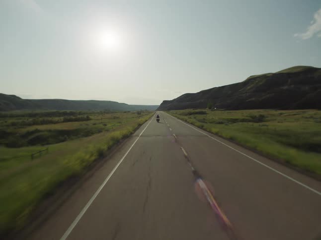 Motorcyclist rides along a long road surrounded by plains and hills under the sun