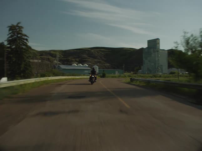 A person riding a motorcycle on a rural road passing a farm making a turn and entering a bridge