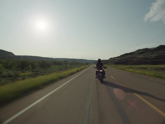 A person riding a motorcycle on a rural road passing hills and grass fields