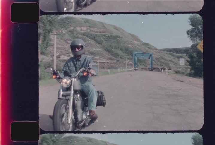 Motorcyclist rides forward on a road flanked by hills on a clear day