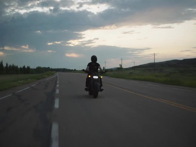 A person wearing a helmet and dark clothes rides a motorcycle directly toward the camera with the headlight on on an empty rural road with fields and rolling hills under a dramatic cloudy sky at dusk