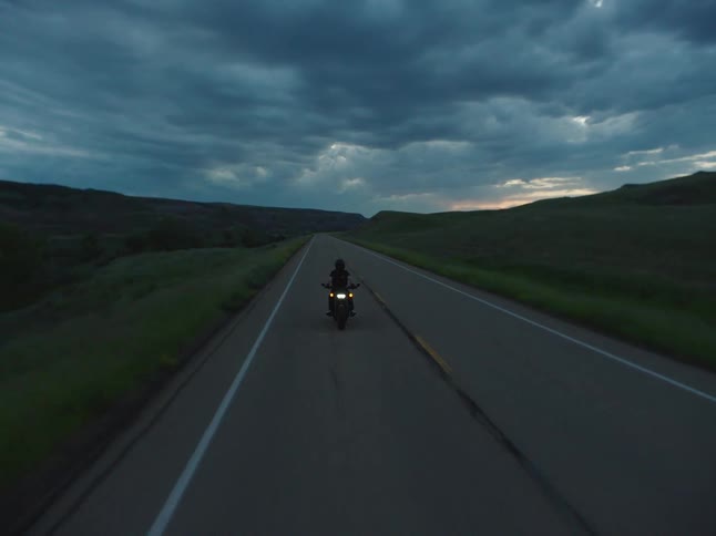 A person riding a motorcycle on a rural road across grass plains and hills under a cloudy sky