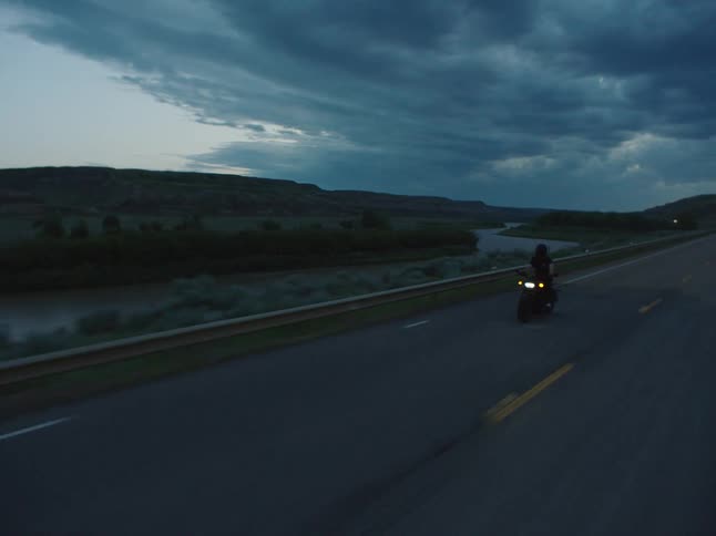Motorcyclist rides along a road beside a river under a cloudy sky at dusk