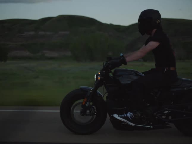 A motorcycle rider man wearing a black helmet and dark shirt drives a dark bike with the headlight on along an empty rural road moving into the foreground under a dramatic dark twilight sky and shadowed hills