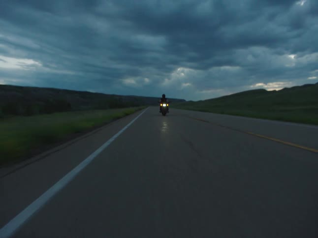 An adult rides a motorcycle on a rural highway at dusk