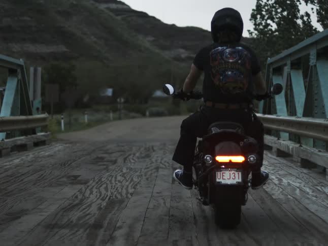 A man wearing a dark tshirt and helmet rides a black motorcycle away from the camera on a wooden and steel bridge at dusk with hills in the background