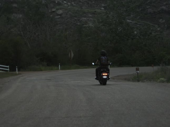 A dark silhouette of a person on a motorcycle with its red tail light glowing rides down a curving dark paved road bordered by a white fence and shadowed hillsides at dusk