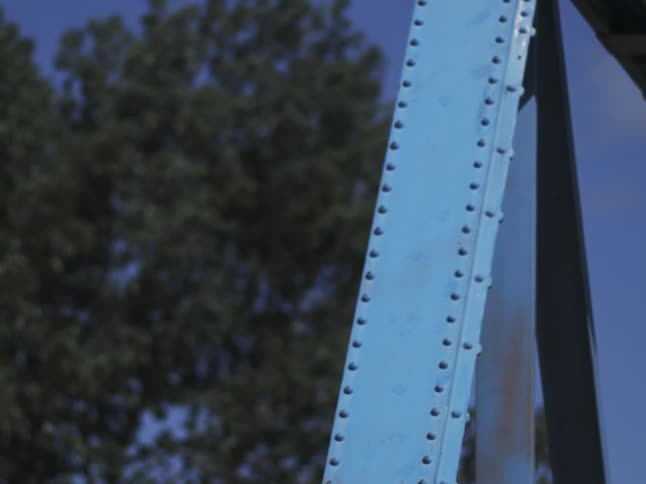 A closeup on paleblue riveted steel beams of a bridge rusty streaks showing against sky and leafy bokeh