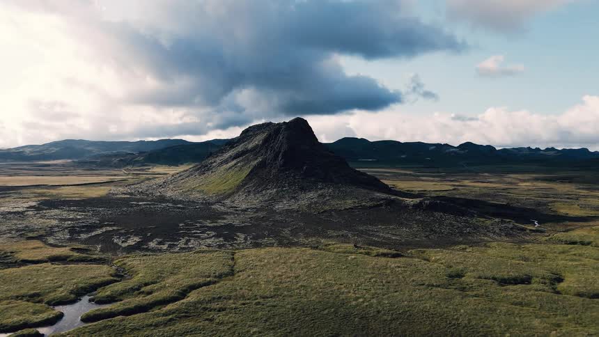 A misty aerial view shows a lone road snaking across dark lava plains toward a low hazy mountain as braided rivers glint under heavy clouds