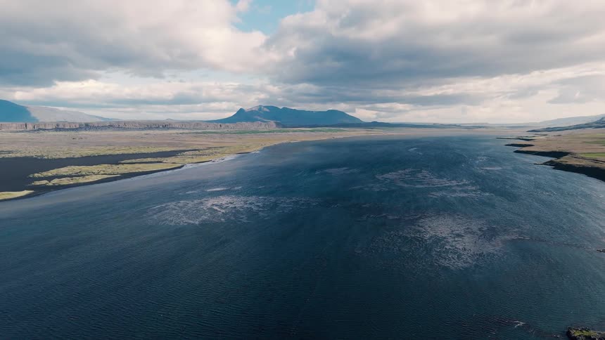 A wide windrippled bay curves between grassy headlands and distant mountains beneath dramatic layered clouds