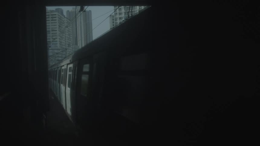 A side profile of a moving railcar emerging from a dark foreground area revealing a densely built collection of tall structures against a pale sky in the background