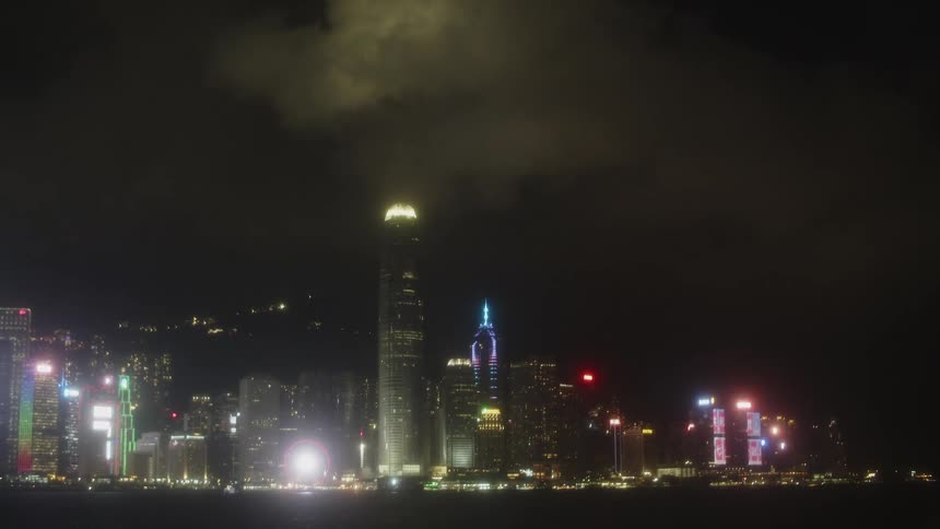 A stationary wide view of a collection of tall structures displaying colored lights at night across a body of water