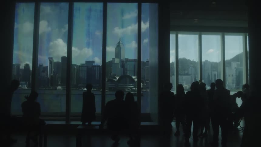 People are silhouetted against large windows while looking across the water towards coastal skyscrapers