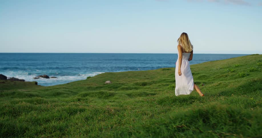 Woman walks alone across a grassy field toward the sea under a blue sky