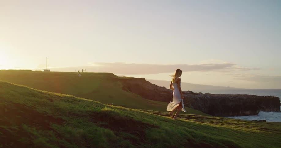 Woman in a white dress walks down a grassy hill at sunset by the sea