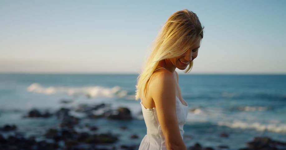 Woman in a white dress looks to the side by the sea under the soft light of dusk
