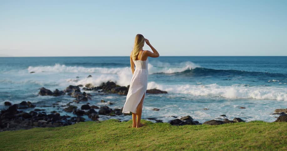 A young blonde woman wearing a long white dress stands barefoot on a grassy cliff edge looking out at blue ocean waves crashing against dark coastal rocks at sunset