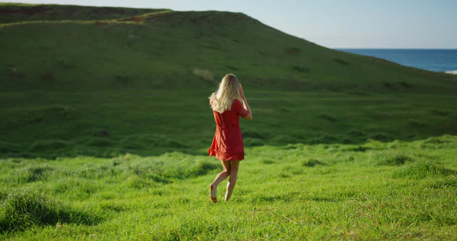 Woman in a red dress walks barefoot across a green field toward the horizon