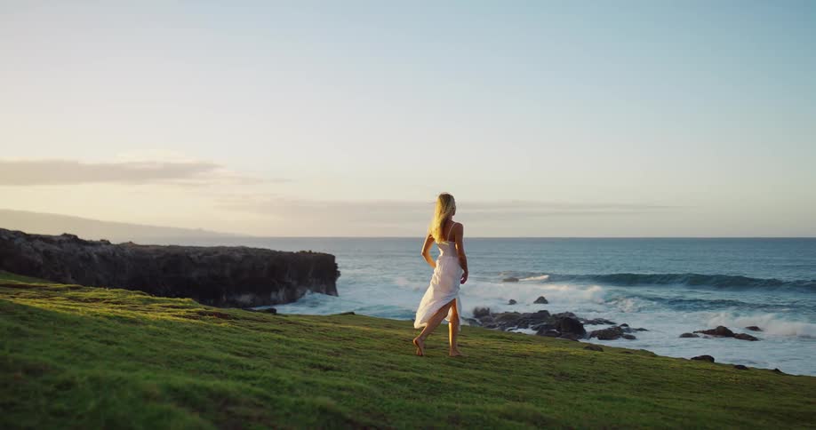 Woman walks barefoot on grassy seaside cliff at sunset wearing a white dress