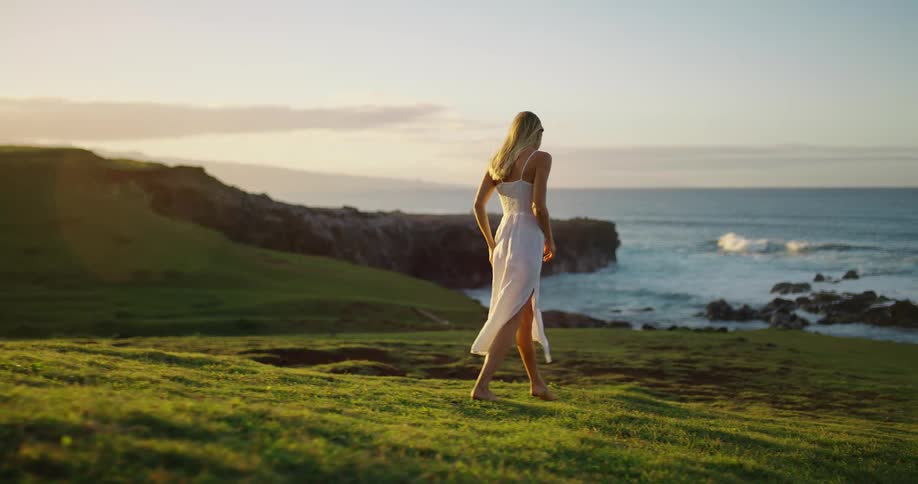 Woman walks barefoot across a grassy hill toward the sea at sunset