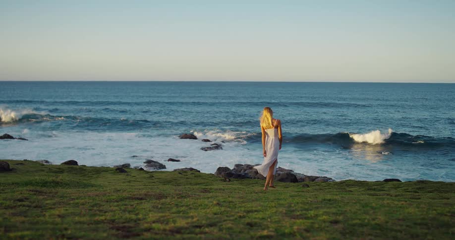 A barefoot blonde woman wearing a white summer dress walks slowly toward the rocky shoreline with crashing blue ocean waves from a grassy cliff edge at sunset