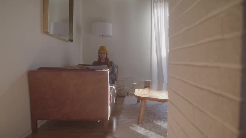 Woman wearing a yellow beanie sits on a sofa lit by the soft morning light
