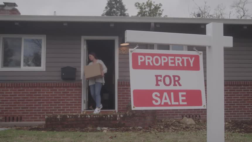 Woman carries a box outside a house with a for sale sign in yard