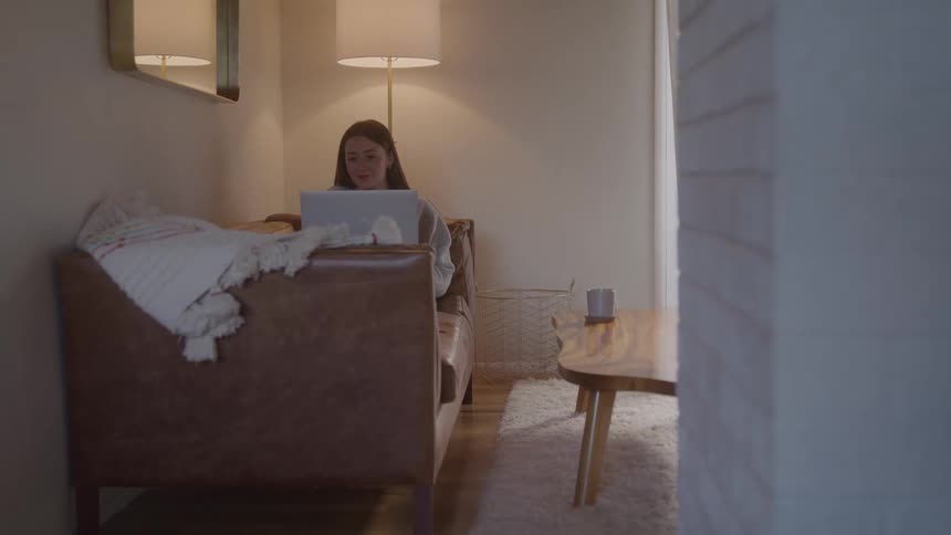A woman sits on a leather sofa working on a laptop beneath a warm floor lamp with a mug on the wooden coffee table and a plush rug softening the cozy room