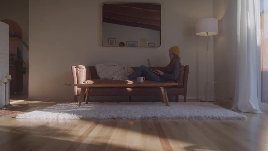 A woman in a yellow beanie lounges on a midcentury couch with a laptop sunlit room around her looking relaxed and focused