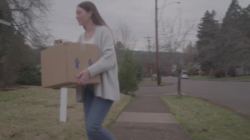 The girl carries a box past the for sale sign in front of the home