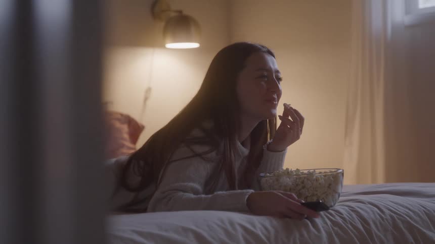 Woman on a bed eating popcorn while watching a tv show in a bedroom