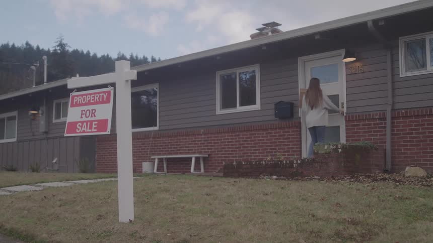 Young woman carrying a cardboard box and entering a house that has a for sale sign in front outdoors during day time