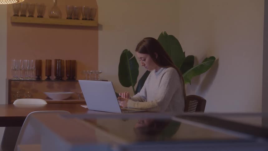 A woman types on a white laptop at a wooden dining table beside a large plant looking concentrated and serious