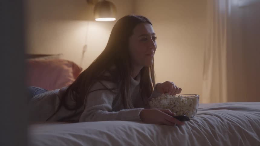A woman lounges on a bed under warm light smiling while eating popcorn and holding a remote looking content