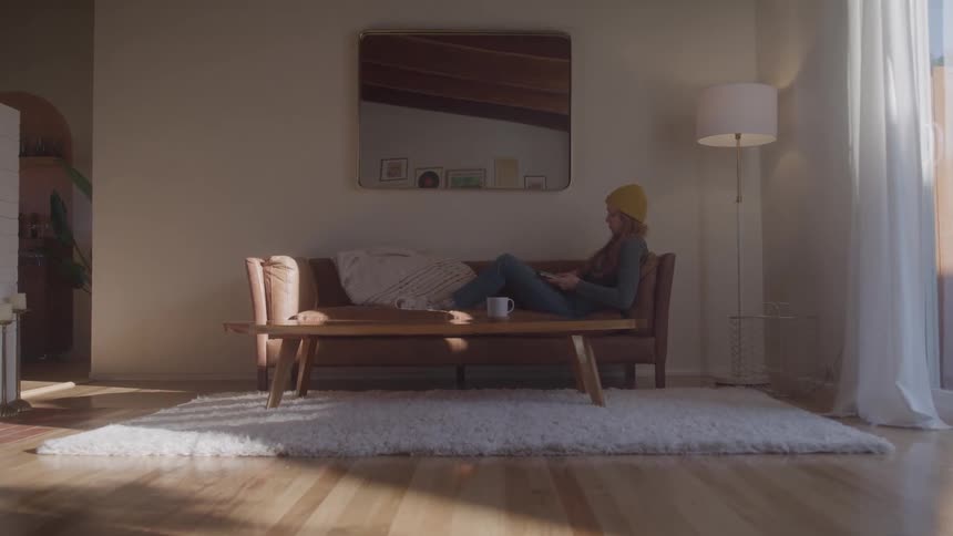 Woman wearing a yellow beanie lying down on a couch on a living room picking up a laptop computer and opening its lid indoors during day time