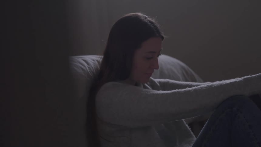 A woman sits in dim light with arms wrapped around her knees gazing ahead looking pensive and sad