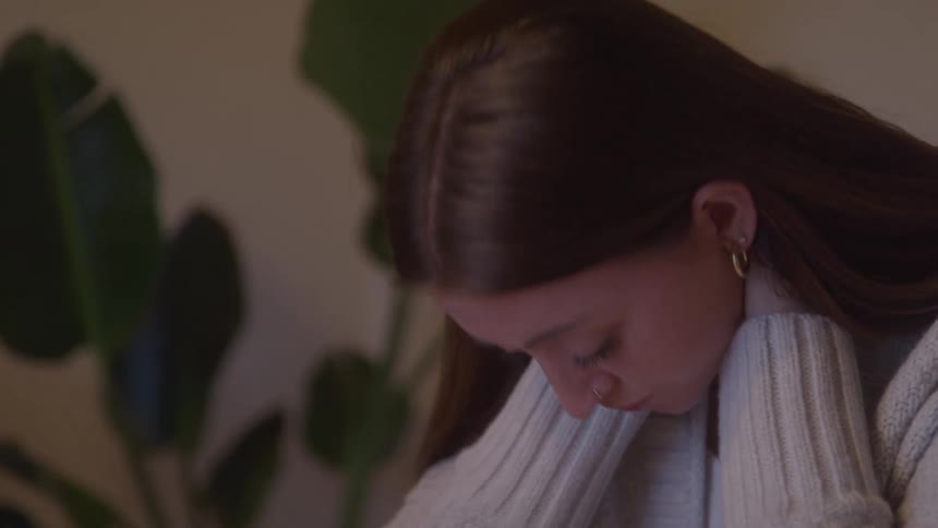 Woman in a light sweater rests her hands on her neck with an introspective expression