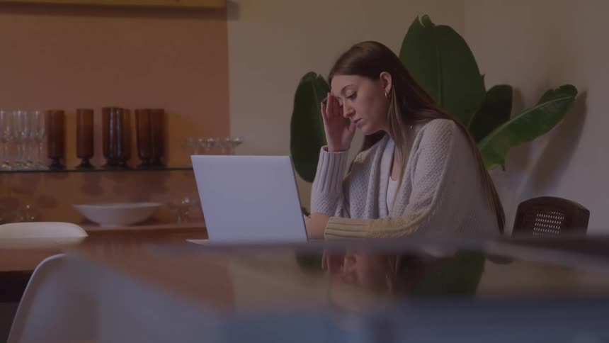 Woman sitting works on laptop with earphones in a welllit home setting