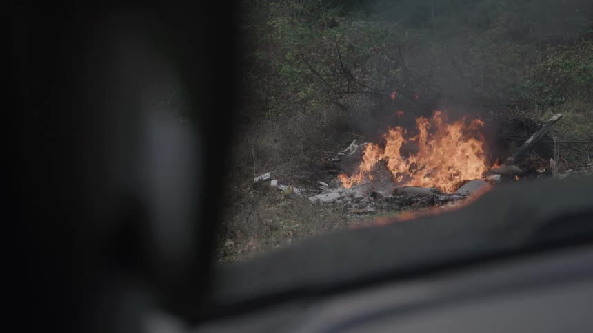 Fire engulfs debris in an open area as seen from inside a vehicle
