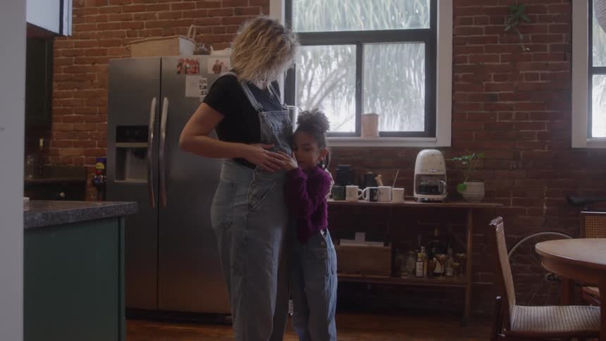 Pregnant mother holds daughters hands in kitchen with brick wall