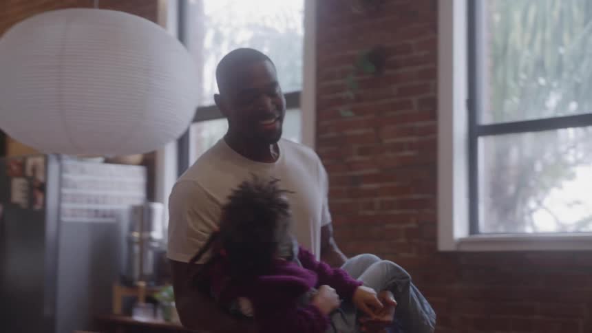Father holds daughter upside down in playful moment in kitchen with brick wall