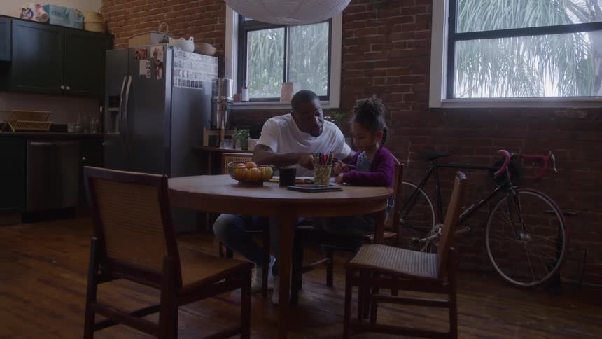 Father helps daughter with activity at round table in room with brick wall