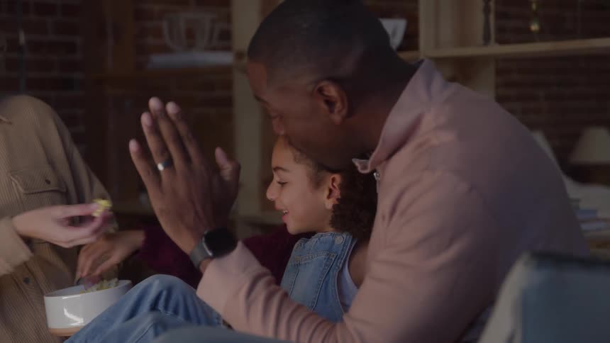 Three people sitting on couch engage with tablet while child eats popcorn